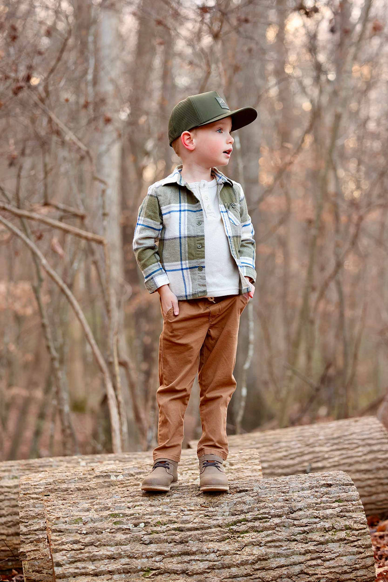 Young boy standing on a log in a forest wearing a Hunter Green colored Rover premium performance fitted hat from Hatchet Headwear, green plaid shirt, brown pants, and brown shoes.