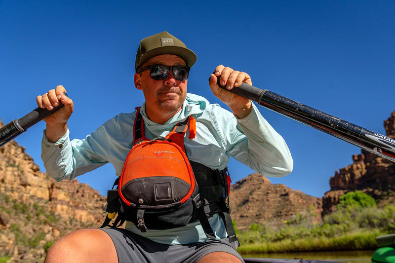 A man wearing a Hunter Green colored Rover premium performance fitted hat from Hatchet Headwear, light blue long-sleeve shirt, sunglasses, and a bright orange and black life vest paddles a kayak on a river with rocky canyon mountains and a clear blue sky in the background.