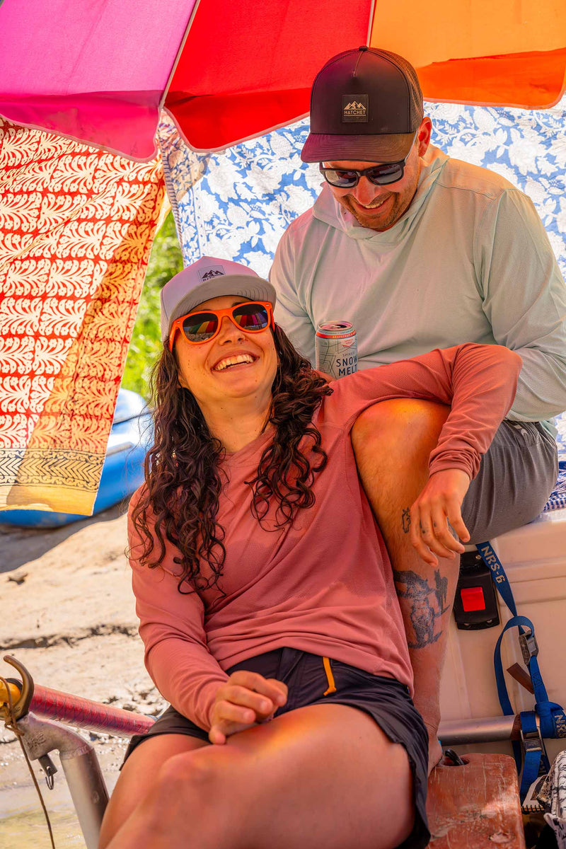 Woman wearing a Glacier colored Rover premium performance fitted hat from Hatchet Headwear, a pink long sleeve shirt, black shorts, and orange sunglasses while leaning/resting against a man wearing a Black colored Rover premium performance hat, light blue long sleeve shirt, and gray shorts.  The couple is sitting on the shore of a lake under a multi-colored umbrella.