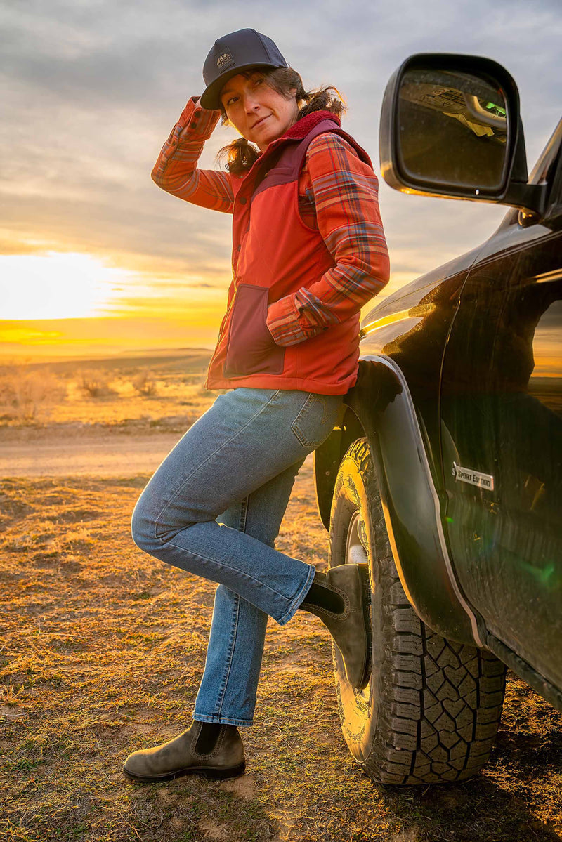 Woman wearing a Slate Blue colored Rover premium performance fitted hat from Hatchet Headwear, an orange plaid shirt, orange vest, and blue jeans while leaning/resting against an SUV with a warm desert sunset in the background.  Woman is facing towards the camera while holding the back of her fitted hat.