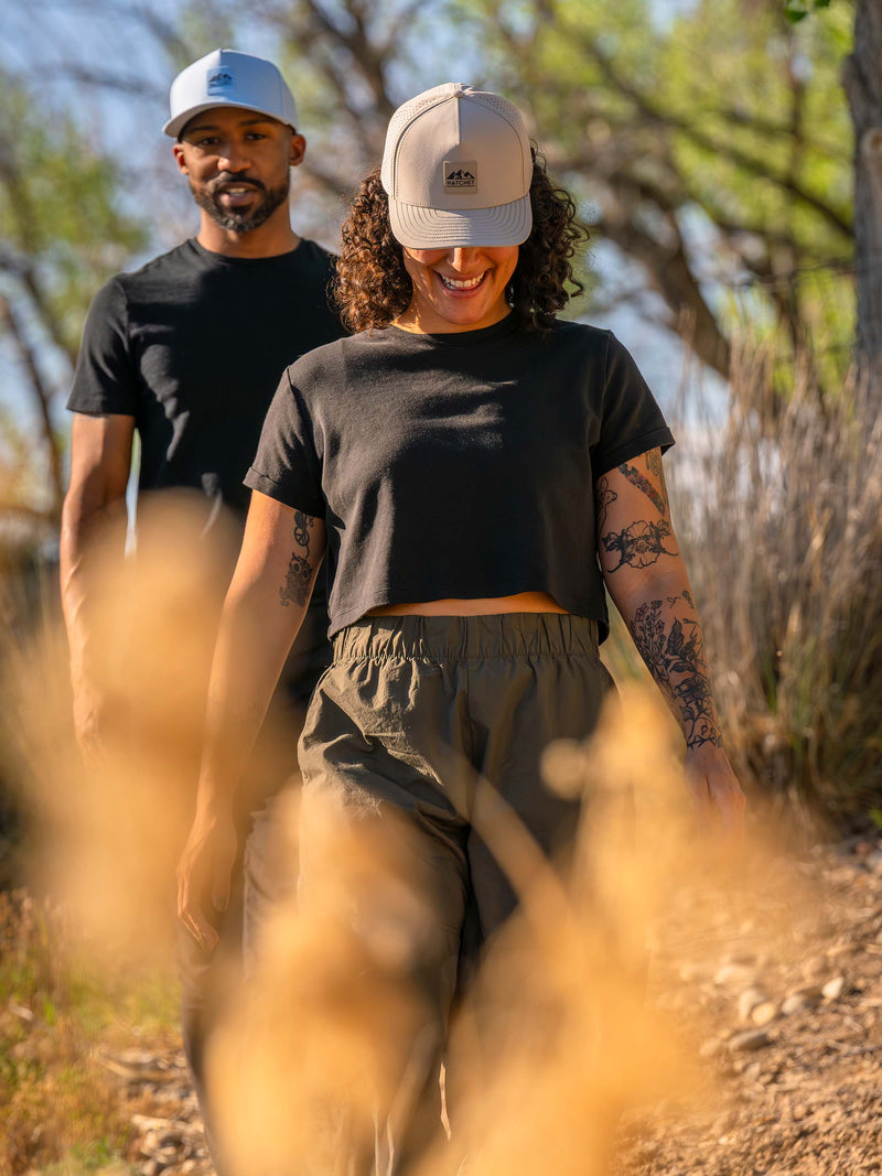 Woman wearing a Boulder colored Rover premium performance fitted hat from Hatchet Headwear, a black shirt, green hiking pants while hiking with a man wearing a Glacier colored Rover premium performance fitted hat, black shirt, and black hiking pants. The couple is hiking through a wooded desert landscape with a warm desert sunset lighting the scene.