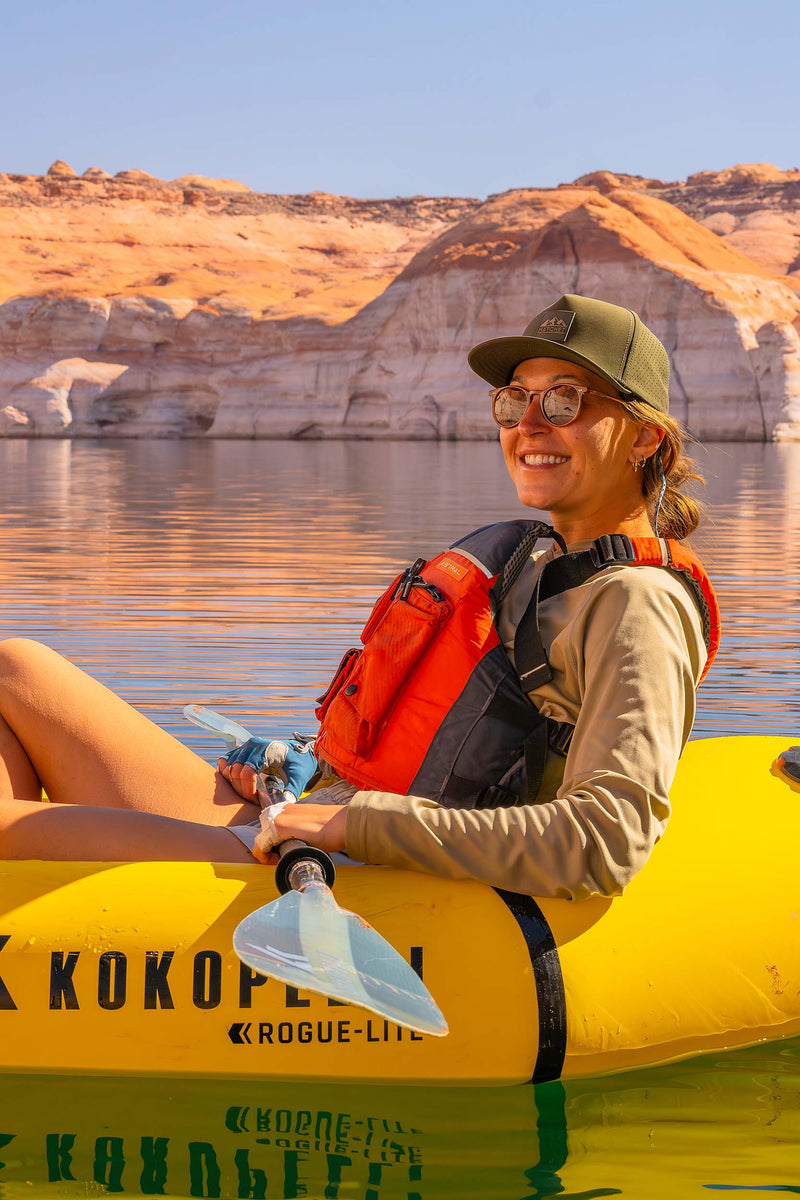 A woman wearing a Hunter Green colored Rover premium performance fitted hat from Hatchet Headwear, sunglasses, and a beige shirt sits in a yellow inflatable raft on a lake, wearing an orange and black life vest. Rock formations are visible in the background under a clear blue sky.