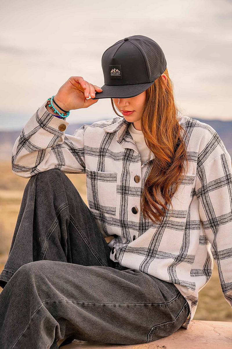 Woman wearing a Black colored Rover premium performance fitted hat from Hatchet Headwear, a black and white plaid shirt, and black jeans while sitting outdoors in front of a scenic desert background.