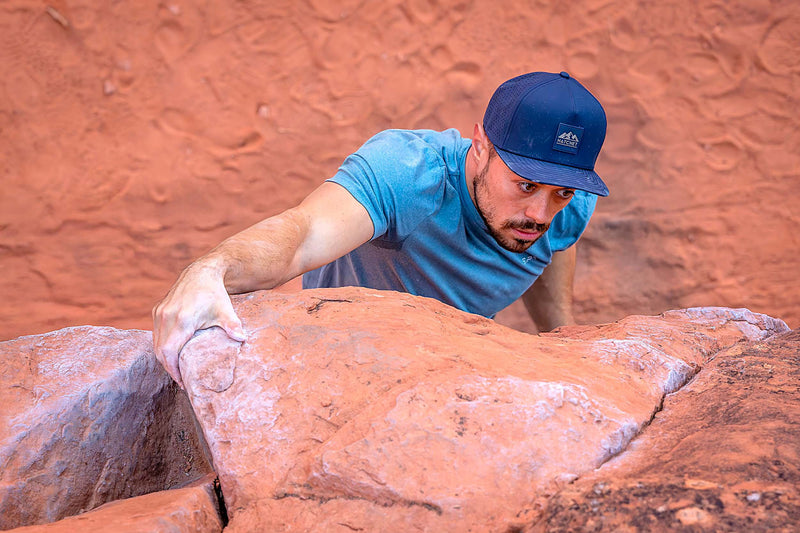 Man climbing a red rock face while wearing a Black colored Rover premium performance fitted hat from Hatchet Headwear and a blue athletic shirt.