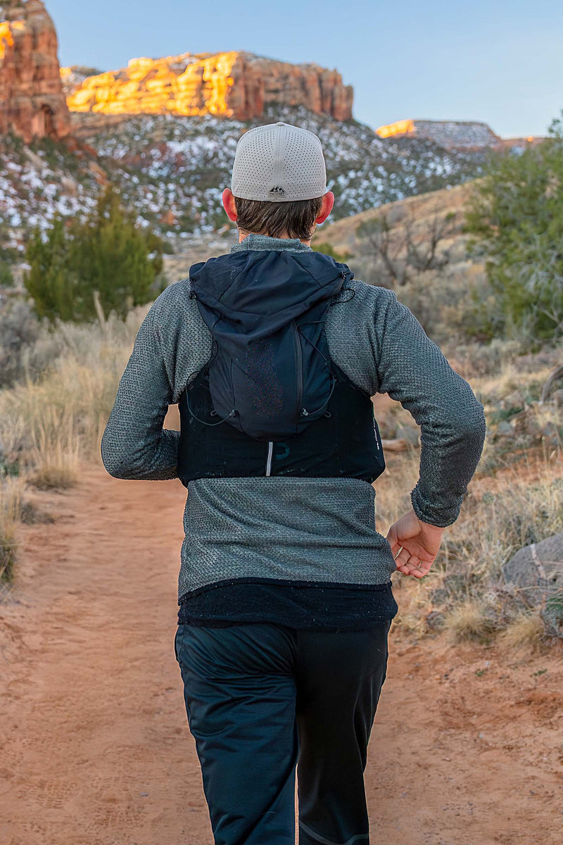 A man running on a desert trail with red rock mountains in the background while wearing a Boulder colored Rover premium performance fitted hat from Hatchet Headwear, a gray waffled long-sleeved shirt, black trail running pants and a hydration vest. 