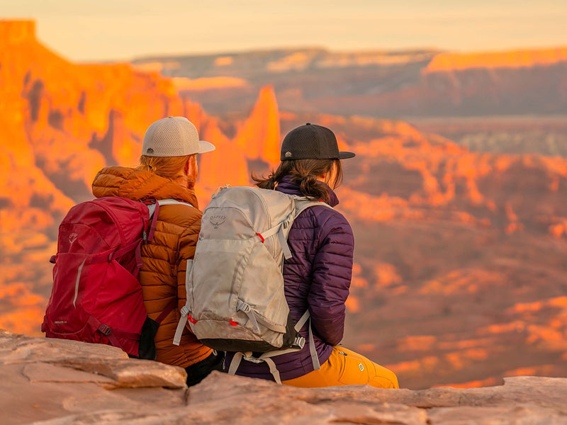 A man and a woman sitting on a rocky outcrop overlooking a red rock canyon at sunset.  The man is wearing a Boulder colored Rover premium performance fitted from Hatchet Headwear, a burnt orange hooded jacket, and a red backpack.  The woman is wearing a Black colored premium performance fitted hat from Hatchet Headwear, a purple jacket, orange pants, and a light gray backpack.