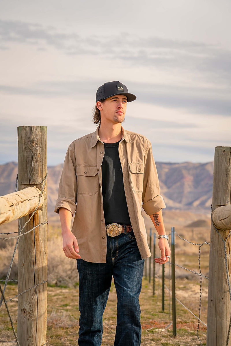 Man walking past a wooden and barbed wire fence while wearing a Black colored premium performance fitted hat from Hatchet Headwear, dark blue jeans, a black undershirt and an unbuttoned tan button-up shirt.  In the background is a mountainous desert landscape.