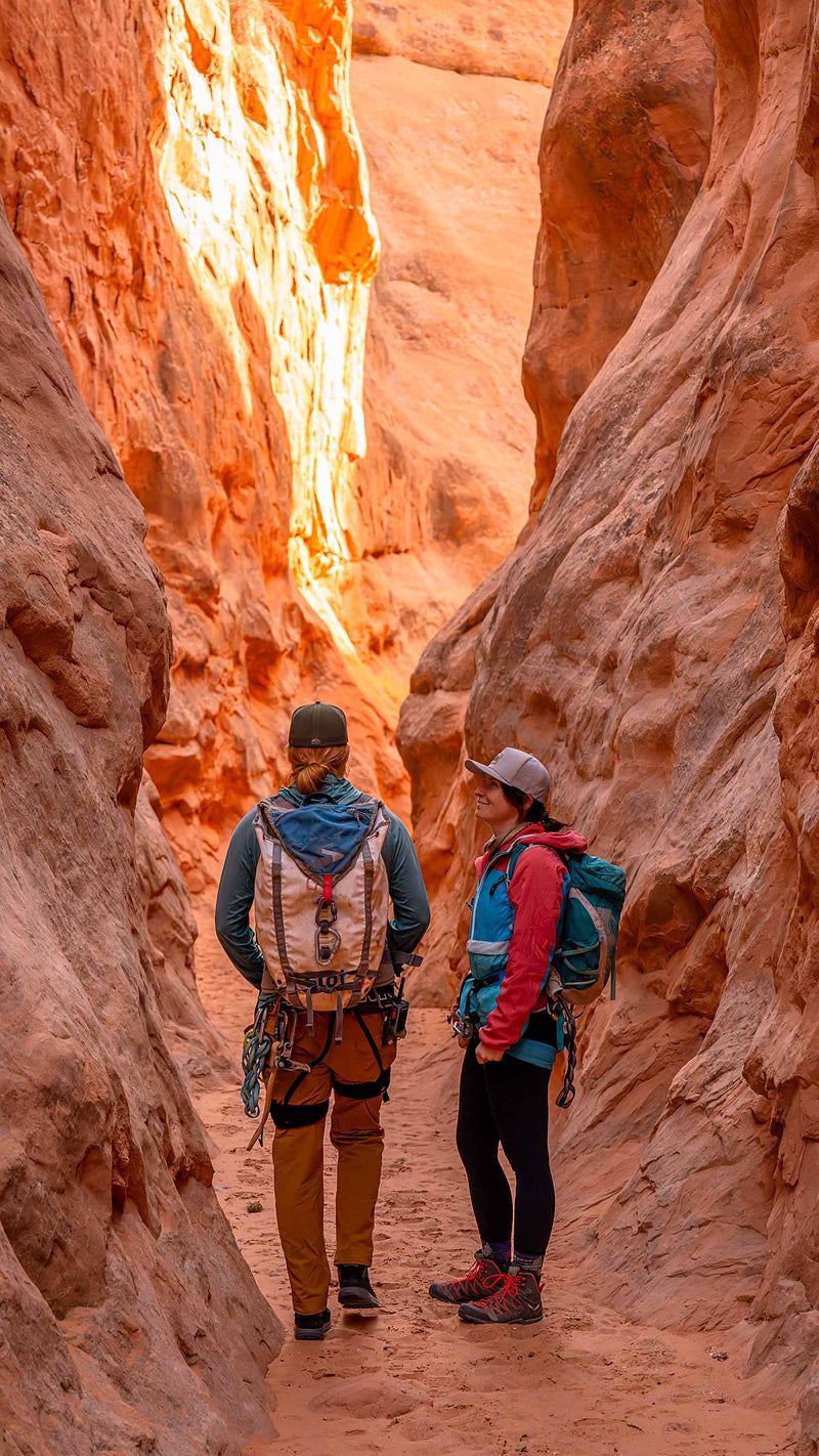Woman wearing a Boulder colored Rover premium performance fitted hat from Hatchet Headwear, teal hiking backpack, red and blue hooded jacket, and outdoor gear while taking a break from hiking through a narrow slot canyon with red rock walls.  The woman is talking to a male hiking partner who is wearing outdoor clothing/gear, a hiking backpack, and a Hunter Green colored Rover premium performance fitted hat from Hatchet Headwear.