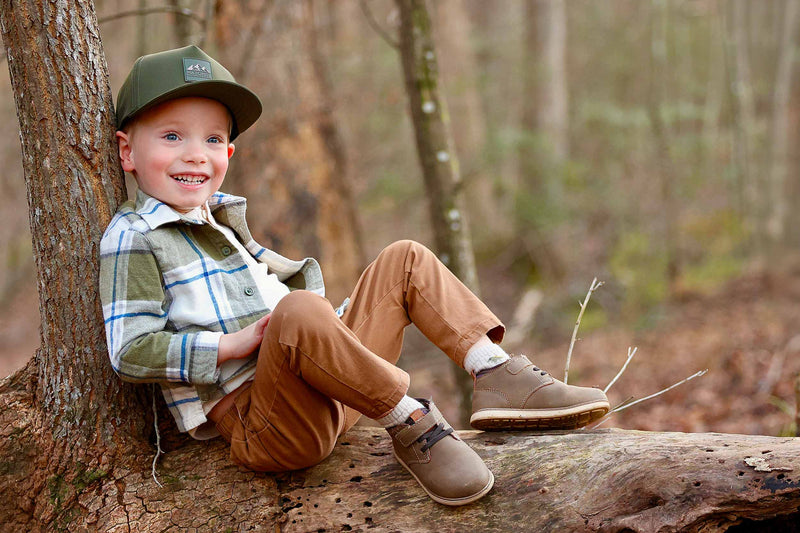 Young boy sitting on a log/leaning against a tree in a forest while wearing a Hunter Green colored Rover premium performance fitted hat from Hatchet Headwear, green plaid shirt, brown pants, and brown shoes.