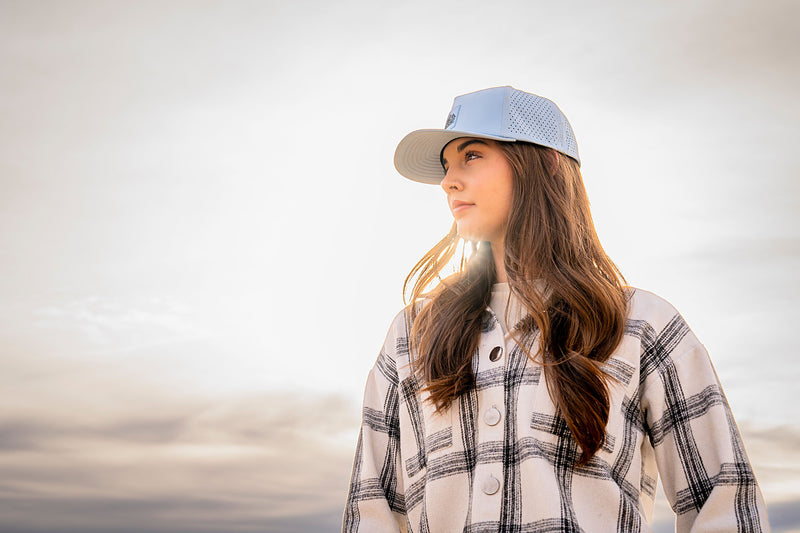 Woman wearing a Glacier colored Rover premium performance fitted hat from Hatchet Headwear and a black and white plaid shirt in front of a scenic sky with bright rays of sun.
