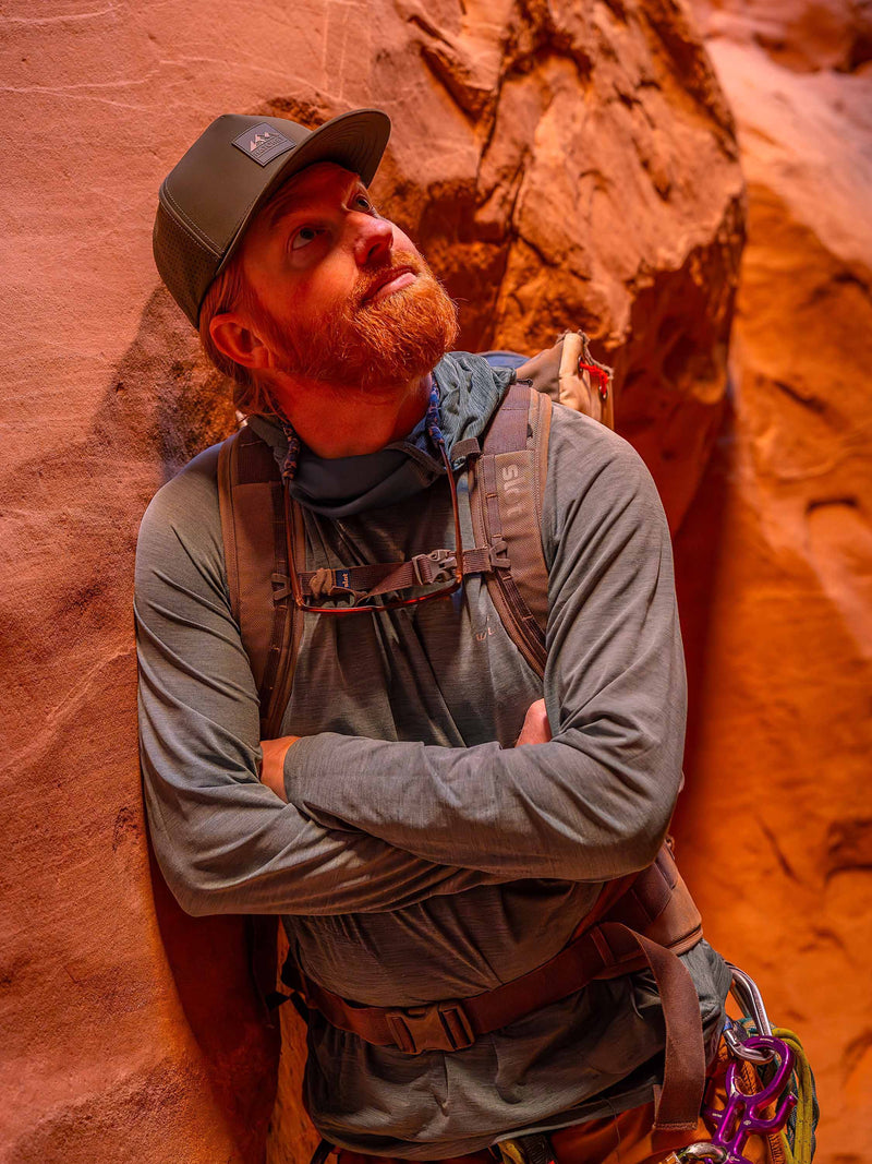 Man wearing a Hunter Green colored Rover premium performance fitted hat from Hatchet Headwear, blue long-sleeve outdoor shirt, gray hiking backpack, and climbing gear. The man is leaning against a red rock canyon wall while looking up through the narrow slot canyon.