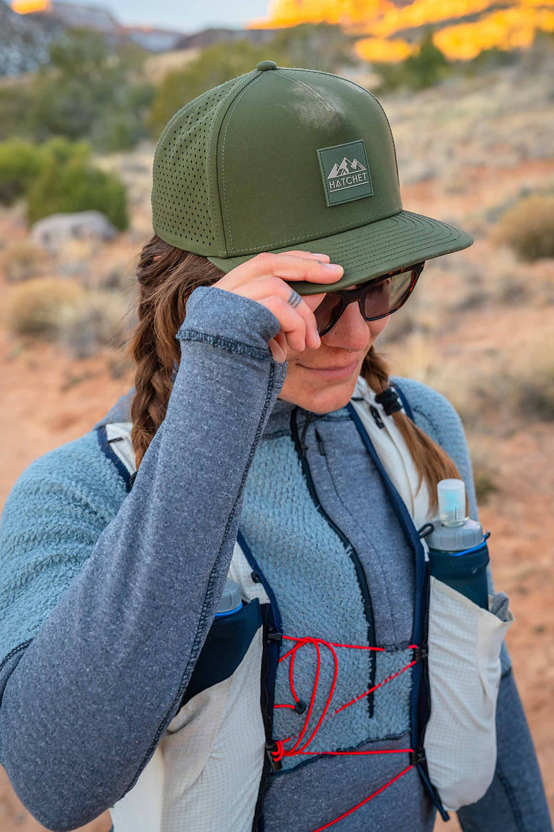 Woman wearing a Hunter Green colored Rover premium performance fitted hat from Hatchet Headwear, sunglasses, a fleece jacket, and a hydration vest. The woman is grabbing the bill of the hat with her right hand while walking through a desert landscape.