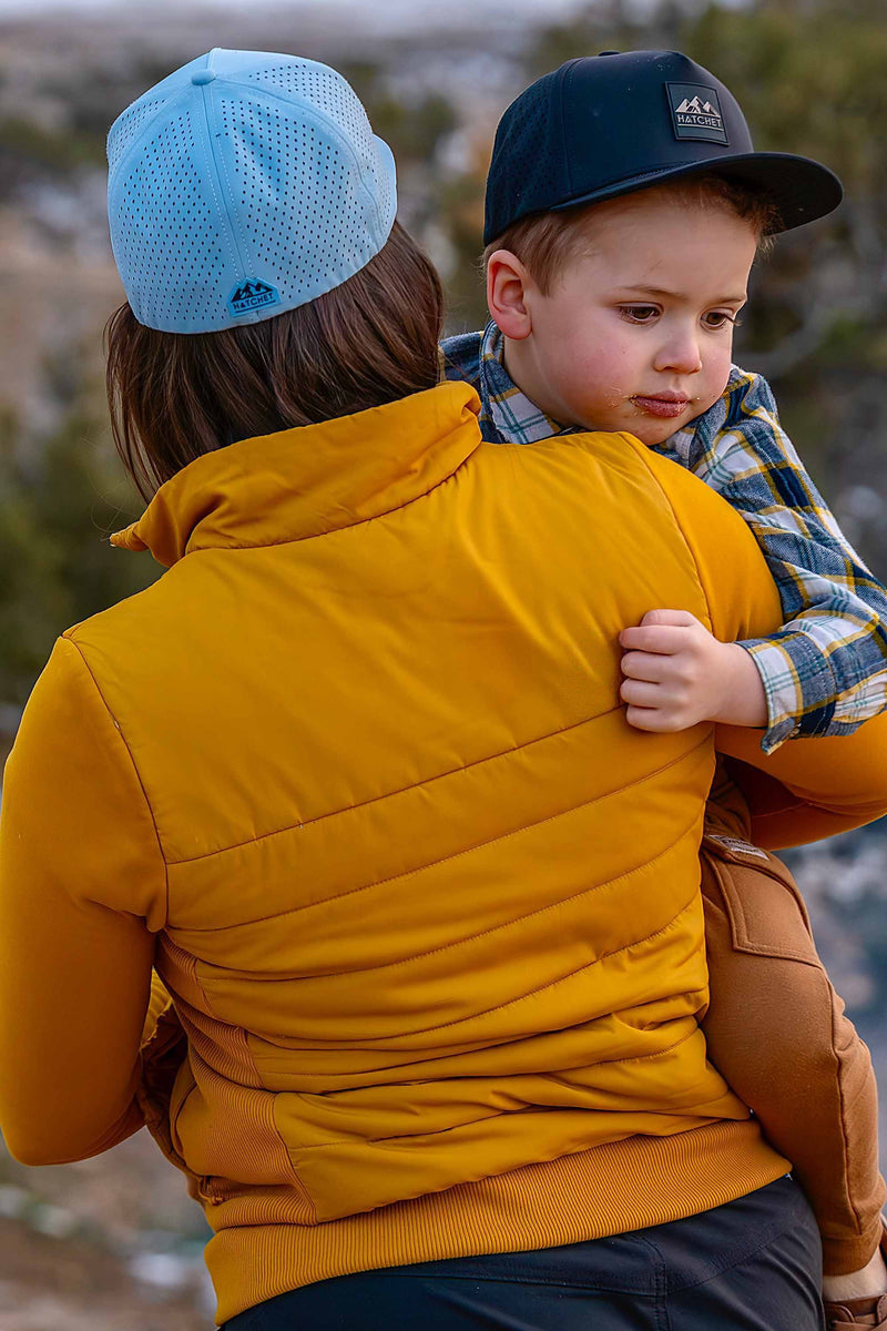 Mother and son wearing Rover premium performance fitted hats from Hatchet Headwear. Mother is wearing a Glacier colored Rover hat, and mustard yellow jacket, and charcoal hiking pants while holding her son in her arms and walking away from the camera through a blurred desert landscape.  Son is looking at the camera and wearing a Black colored Rover hat, brown cargo pants, and a blue, white and yellow plaid shirt.