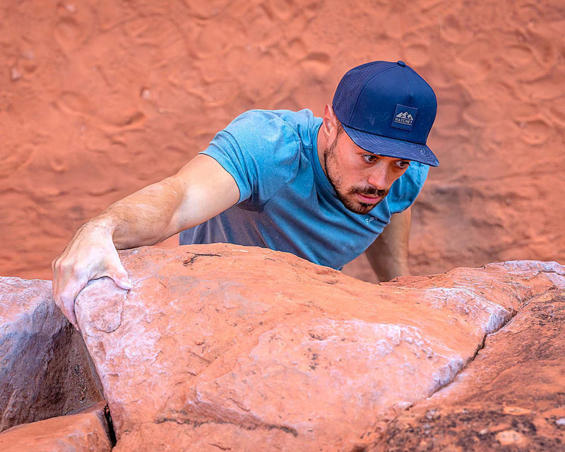 Man climbing a red rock face while wearing a Black colored Rover premium performance fitted hat from Hatchet Headwear and a blue athletic shirt.