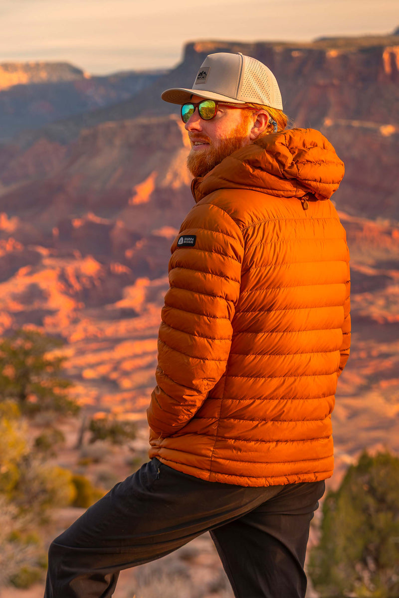Man wearing a Boulder colored Rover premium performance fitted hat from Hatchet Headwear, sunglasses, an orange puffer jacket, and brown hiking pants. The man is looking off into the distance while standing in front of a  blurred desert canyon landscape.