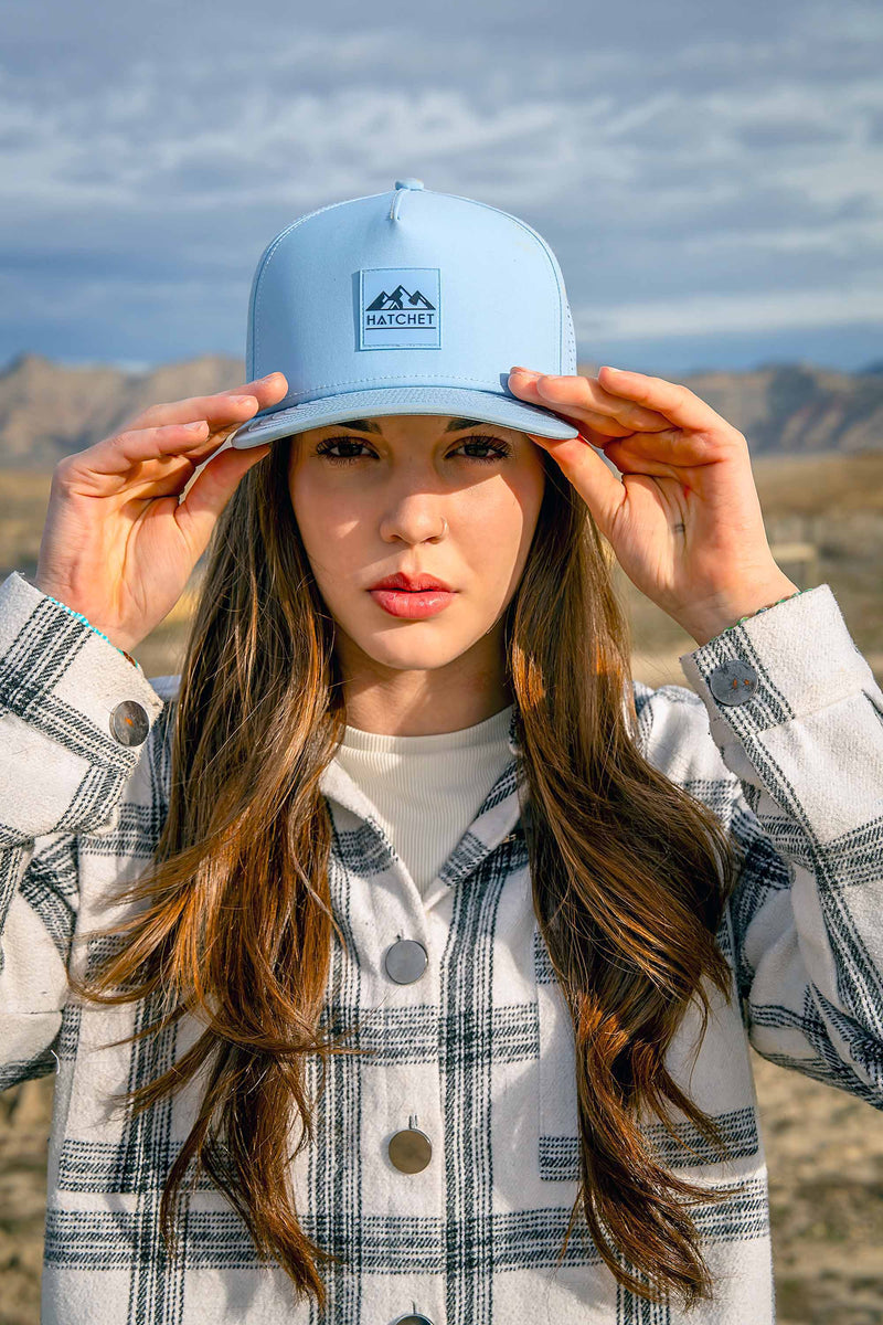 Young woman wearing a Glacier colored Rover premium performance fitted hat from Hatchet Headwear and a black and white paid shirt. The woman is grabbing the bill of the hat with both hand while standing in a desert landscape.