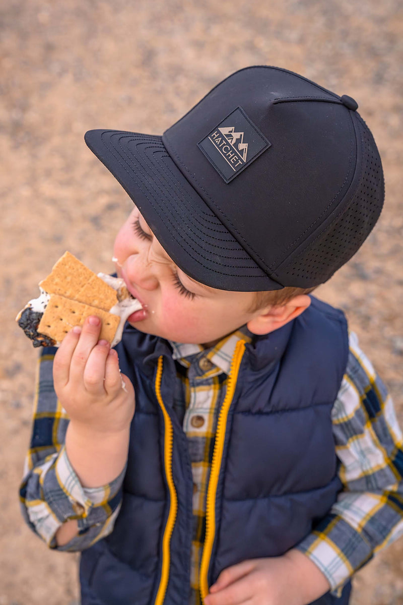 Young boy wearing a Black colored Rover premium performance fitted hat from Hatchet Headwear, a blue vest, and a blue, white and yellow plaid shirt while eating a s'more outdoors.