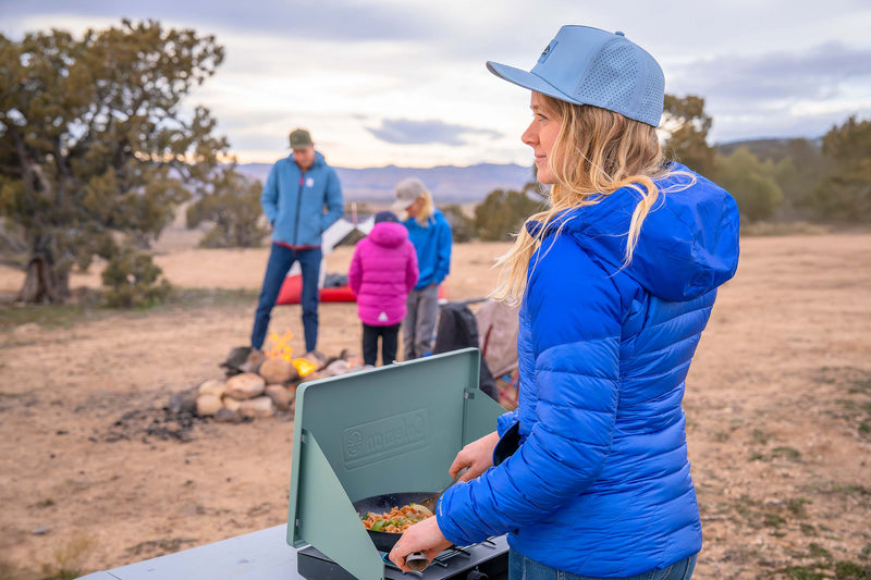 Woman wearing a Glacier colored Rover premium performance fitted hat from Hatchet Headwear, blue puffer jacket, and blue jeans while cooking on a portable camping grill.  In the blurred desert landscape background, a man and two children wearing outdoor clothing/gear and Rover premium performance fitted hats from Hatchet Headwear are visible.