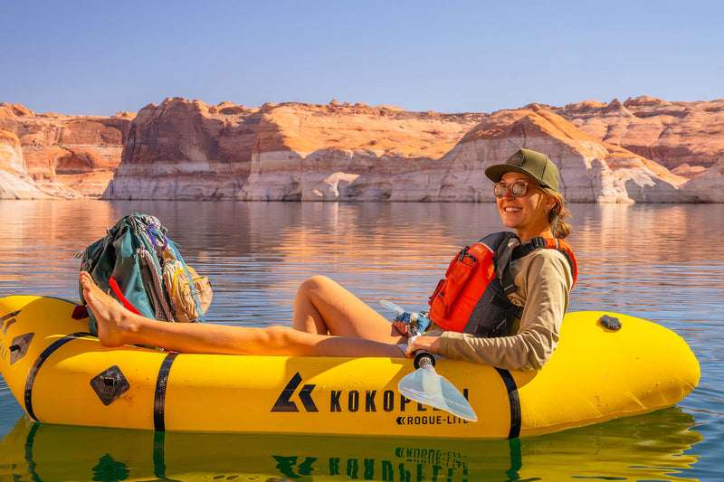 A woman wearing a Hunter Green colored Rover premium performance fitted hat from Hatchet Headwear, sunglasses, and a beige shirt sits in a yellow inflatable raft on a lake, wearing an orange and black life vest. Rock formations are visible in the background under a clear blue sky.