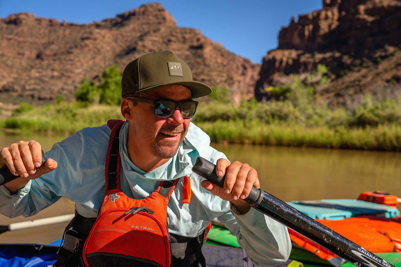 A man wearing a Hunter Green colored Rover premium performance fitted hat from Hatchet Headwear, light blue long-sleeve shirt, sunglasses, and a bright orange and black life vest paddles a kayak on a river with rocky canyon mountains and a clear blue sky in the background.