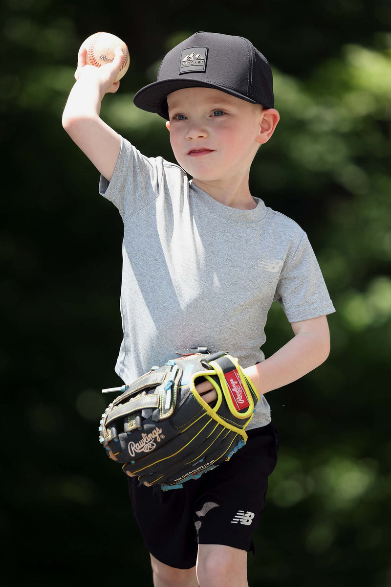 A young boy wearing a Black colored Rover premium performance fitted hat from Hatchet Headwear, gray athletic t-shirt, and black athletic shorts while playing baseball.