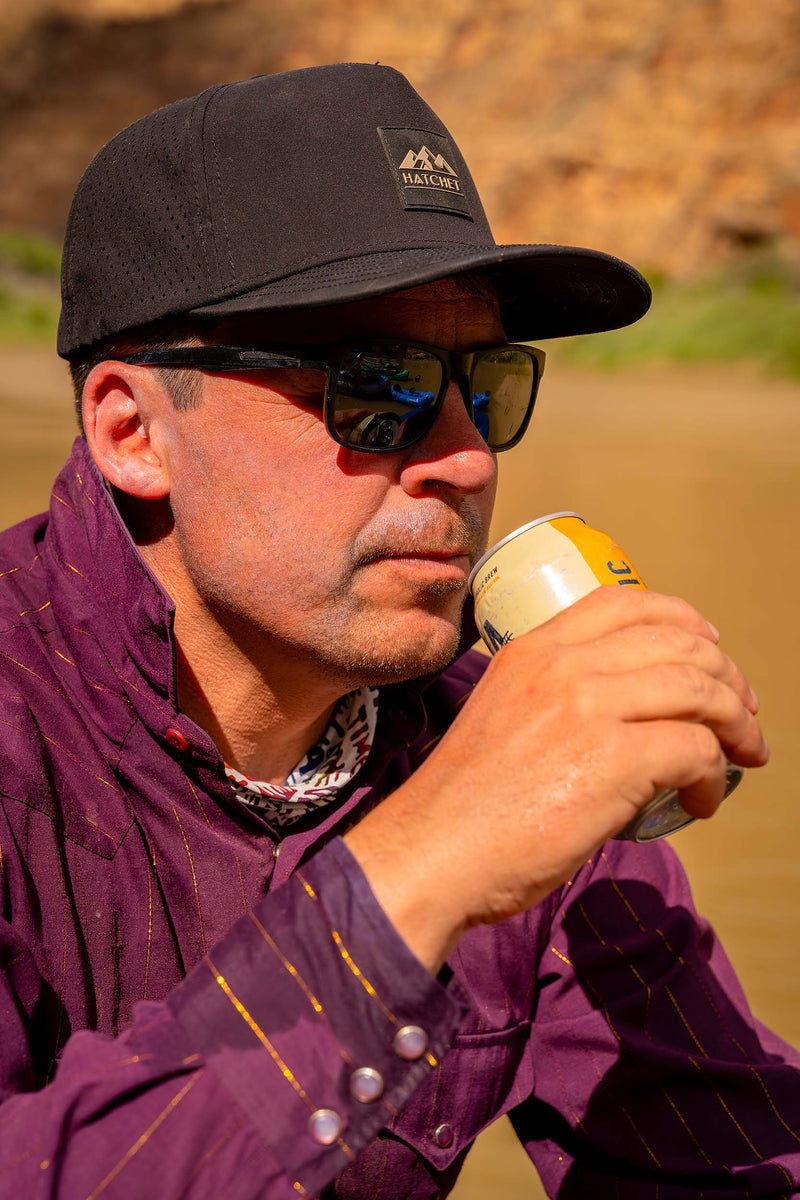 A man wearing a Black colored Rover premium performance fitted hat from Hatchet Headwear, black sunglasses, and a purple striped shirt drinking a canned beverage while sitting on the shore of a river surrounded by red rock canyon walls.