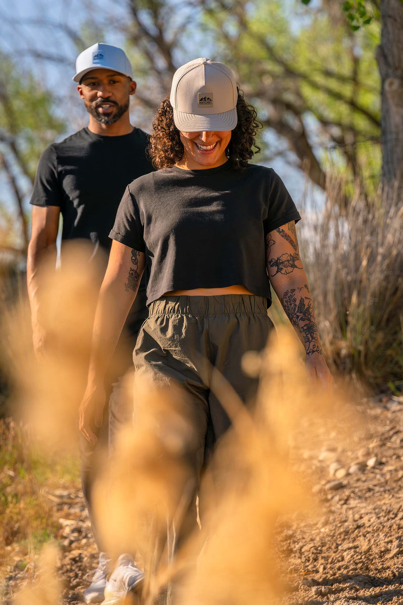 Woman wearing a Boulder colored Rover premium performance fitted hat from Hatchet Headwear, a black shirt, green hiking pants while hiking with a man wearing a Glacier colored Rover premium performance fitted hat, black shirt, and black hiking pants. The couple is hiking through a wooded desert landscape with a warm desert sunset lighting the scene.