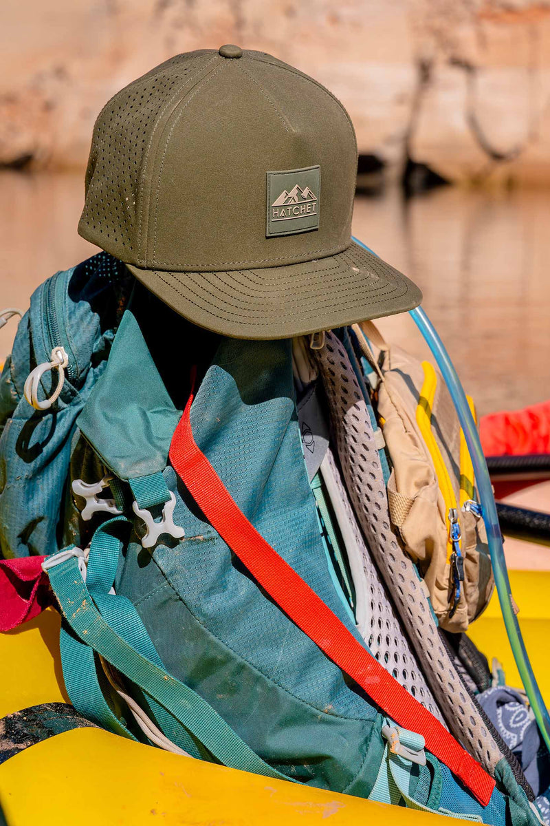 Hunter Green colored Rover premium performance fitted hat from Hatchet Headwear laying on top of a teal hiking backpack that is inside of a yellow kayak. The kayak, hiking gear, and hat are floating down a river in a red rock canyon.