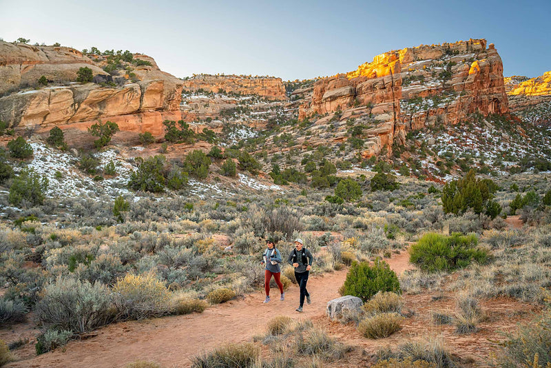 A man and woman running on a desert trail with red rock mountains in the background.  The man is wearing a Boulder colored Rover premium performance fitted hat from Hatchet Headwear, a gray waffled long-sleeved shirt, black trail running pants and a hydration vest.  The woman is wearing a Hunter Green colored Rover premium performance fitted hat from Hatchet Headwear, a grey long sleeved shirt, burnt orange leggings and a hydration vest.