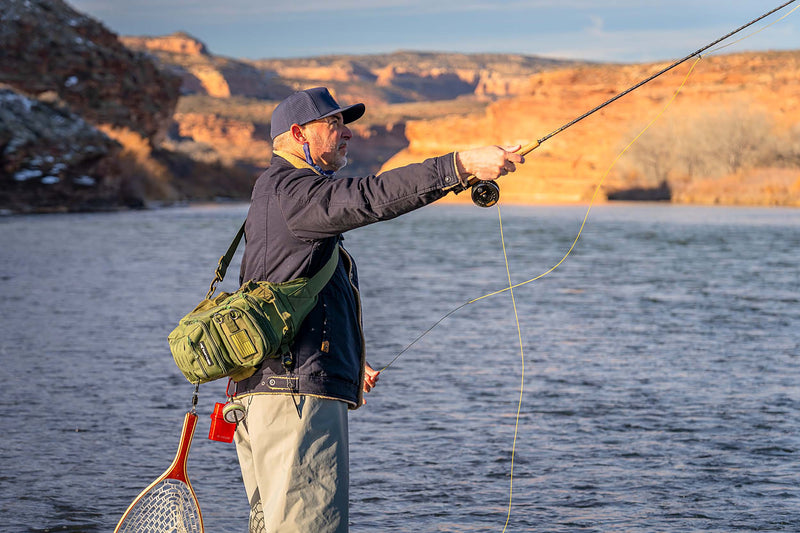 Man wearing a Slate Blue colored Rover premium performance fitted hat from Hatchet Headwear, fleece lined denim jacket, fly fishing bibs, and fly fishing gear while fly fishing in a lake in front of a red rock desert landscape.