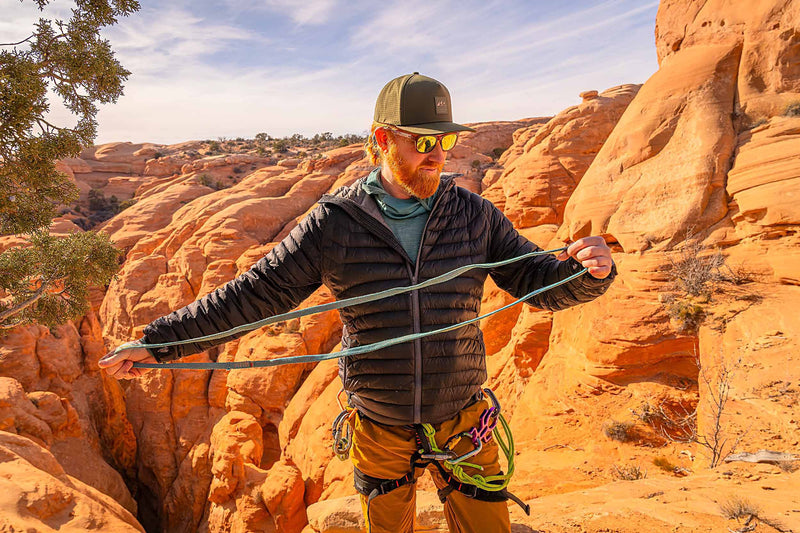 Man wearing a Hunter Green colored Rover premium performance fitted hat from Hatchet Headwear, blue jacket, tan pants, and outdoor gear with climbing equipment in a desert landscape with red rock canyons in the background.